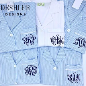 May include: Blue and white striped pajama sets with personalized monogrammed pockets. One white pajama set is also visible. The brand name "Deshler Designs" is printed on a tag. The pajamas have white buttons and trim.