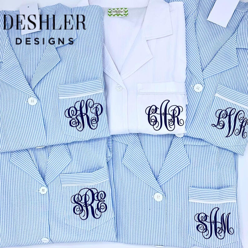 May include: Blue and white striped pajama sets with personalized monogrammed pockets. One white pajama set is also visible. The brand name "Deshler Designs" is printed on a tag. The pajamas have white buttons and trim.