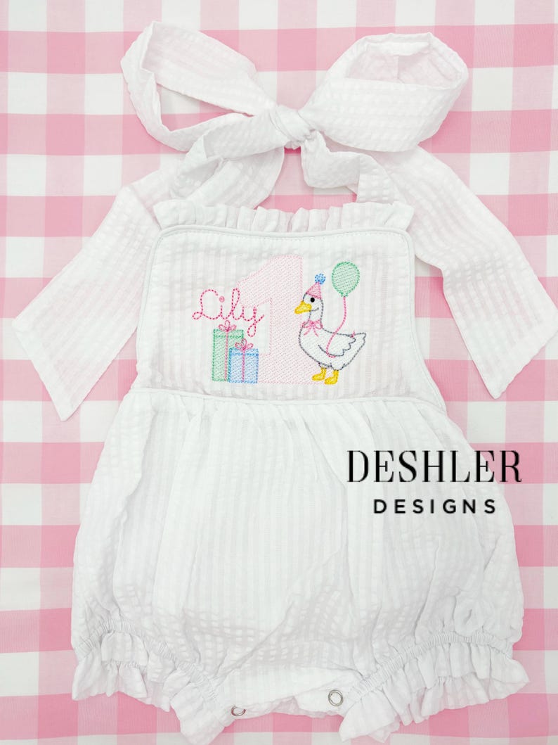 May include: White seersucker romper with a large bow and ruffle trim. The front features an embroidered duck with a party hat and balloon, the name "Lily", and the number "1". The text "DESHLER DESIGNS" is printed below the duck.