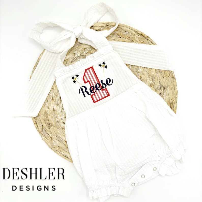 May include: White sleeveless onesie with red and blue stripes and a red and blue number one with the name Reese embroidered on it. The onesie has a bow tie and is on a woven straw background.