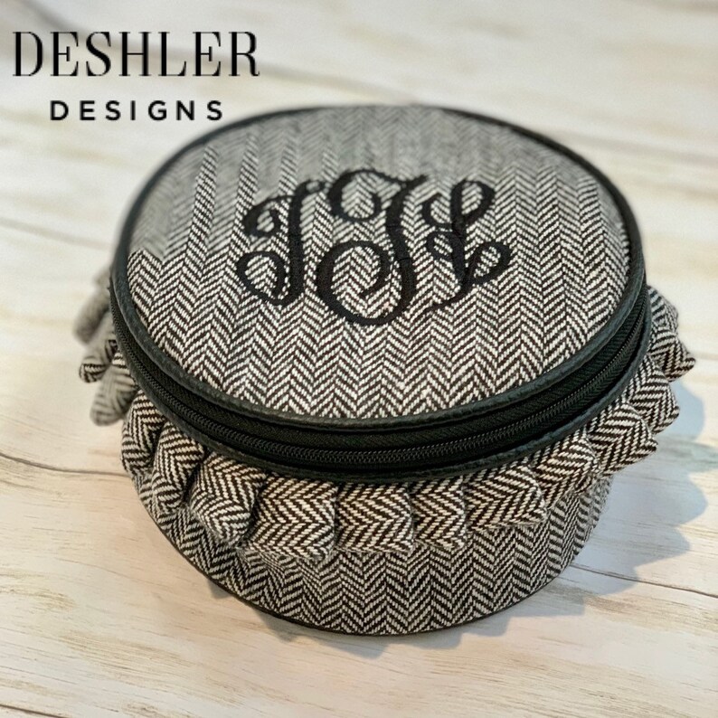 May include: A round, black and white, zippered, fabric jewelry box with a ruffled edge. The box has a monogram of the letters "J.L." embroidered on the top.