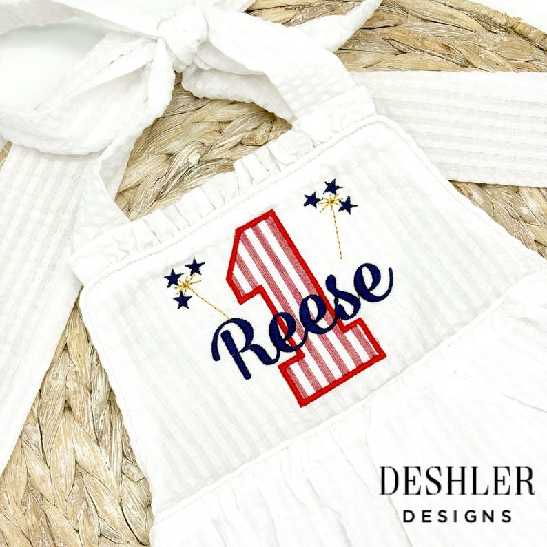 May include: A white sleeveless dress with red and blue stripes and a large red and blue number one with the name Reese embroidered in blue. The dress has a ruffled neckline and is decorated with gold glitter fireworks.