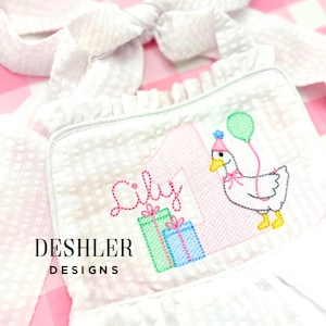 May include: White, textured fabric bib with a pink embroidered number 1, the name "Lily", and a cartoon goose holding a balloon. The bib also features two embroidered gift boxes and the text "DESHLER DESIGNS".