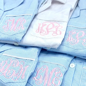 May include: Close-up of several blue and white striped pajama tops with pink embroidered monograms on the pocket. One white pajama top is also visible. The brand name "DESHLER DESIGNS" is printed at the bottom.