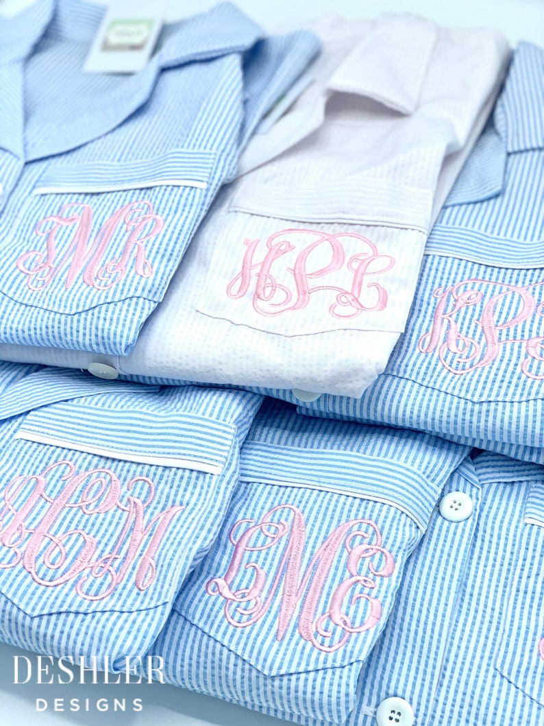 May include: Close-up of several blue and white striped pajama tops with pink embroidered monograms on the pocket. One white pajama top is also visible. The brand name "DESHLER DESIGNS" is printed at the bottom.