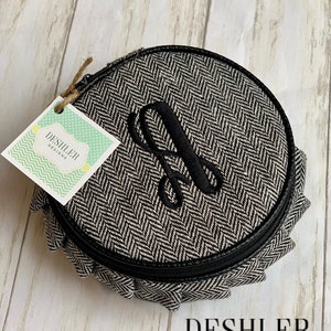 May include: A round, black and white, zippered, fabric pouch with a herringbone pattern. The pouch has a black leather trim and a black embroidered letter "A" on the front. The pouch is sitting on a light wood surface. The text "DESHLER DESIGNS" is visible in the bottom right corner of the image.