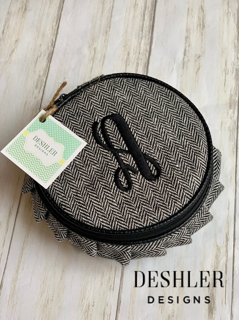 May include: A round, black and white, zippered, fabric pouch with a herringbone pattern. The pouch has a black leather trim and a black embroidered letter "A" on the front. The pouch is sitting on a light wood surface. The text "DESHLER DESIGNS" is visible in the bottom right corner of the image.