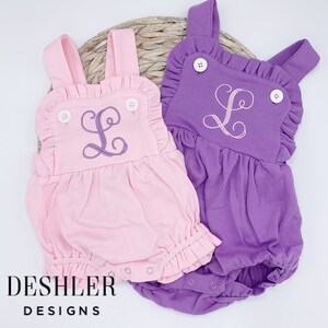 May include: Two baby rompers, one light pink and one purple, each with a ruffled bib and straps. The rompers feature an embroidered letter "L" and white buttons. The brand name "DESHLER DESIGNS" is printed in black.