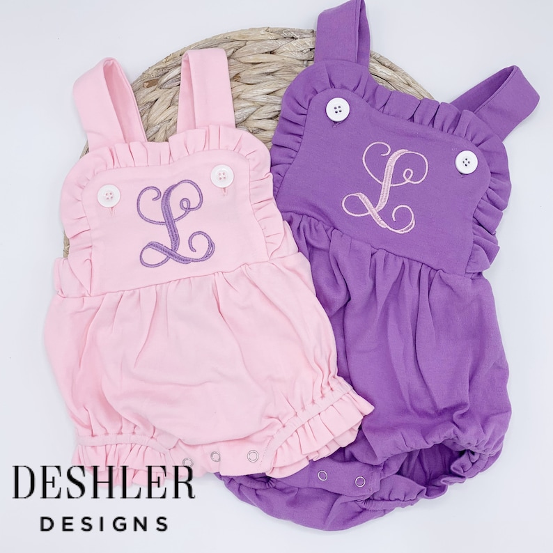 May include: Two baby rompers, one light pink and one purple, each with a ruffled bib and straps. The rompers feature an embroidered letter "L" and white buttons. The brand name "DESHLER DESIGNS" is printed in black.