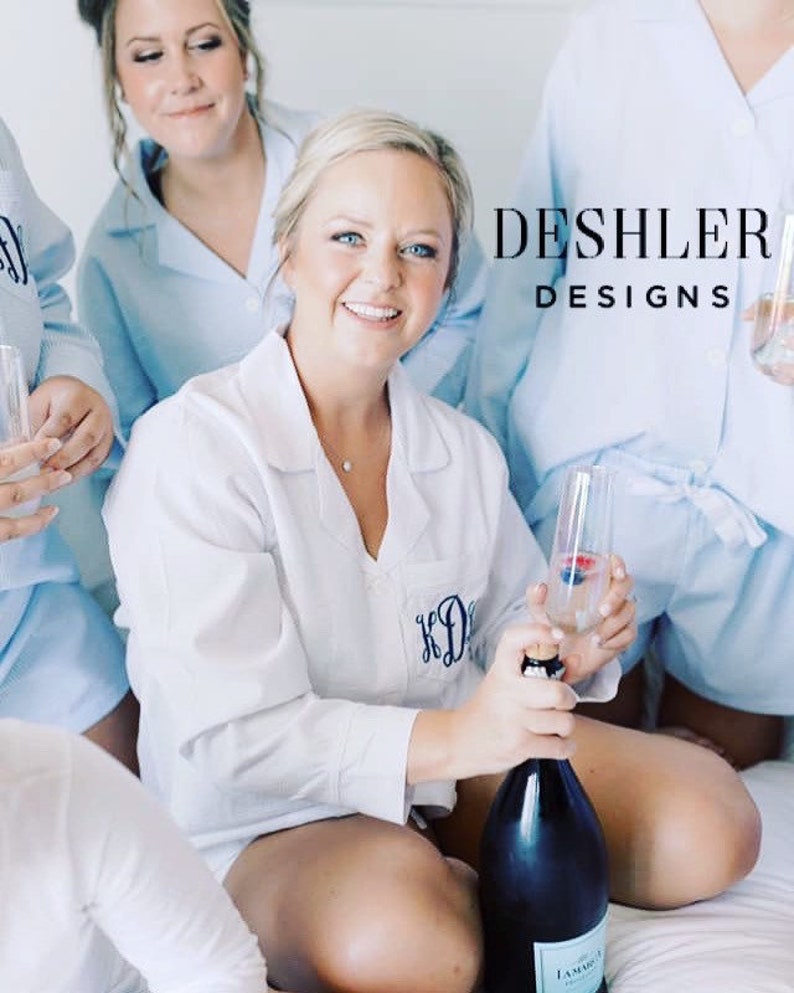 May include: White pajama shirt with a monogram and light blue pajama shorts. A woman is holding a champagne bottle and a glass of champagne. The text "DESHLER DESIGNS" is visible.