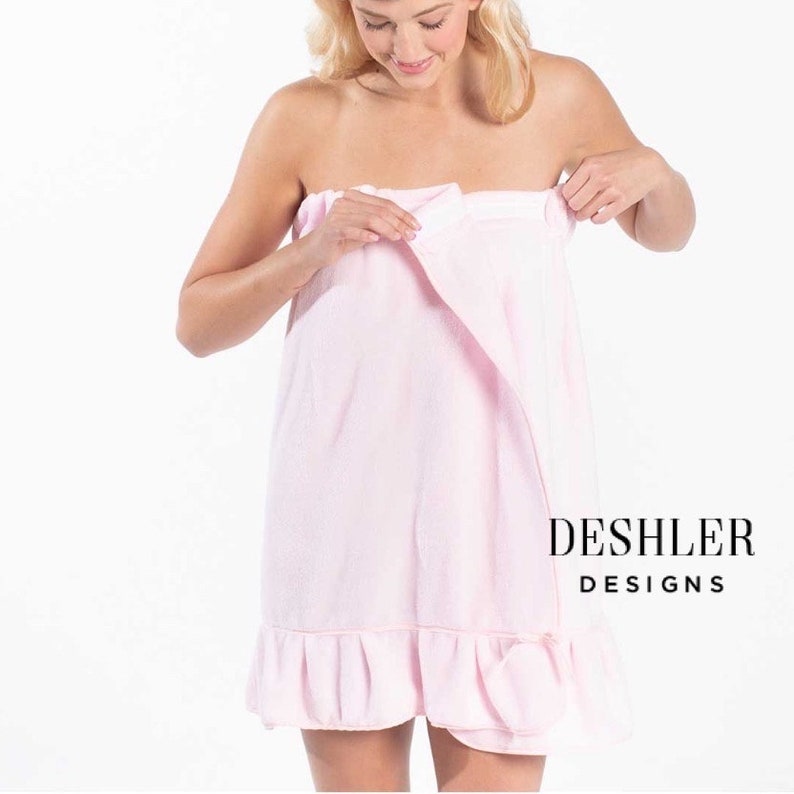May include: A light pink spa wrap with a ruffled hem. The wrap is being secured around the body. The text "DESHLER DESIGNS" is printed in black on the lower right corner. The wrap appears to be made of a soft, absorbent material.