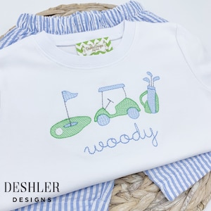 May include: White t-shirt with embroidered golf-themed design. The design includes a golf cart, golf clubs, a golf ball, and a flag. The text "woody" is embroidered below the design.