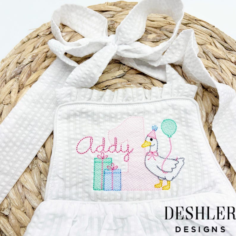 May include: White seersucker romper with a ruffle trim and tie straps. The front features an embroidered design with the name "Addy", a number "1", a goose wearing a party hat, and gift boxes. A balloon is also included in the design.