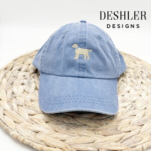 May include: A blue denim baseball cap with a light brown embroidered dog silhouette. The cap has a curved brim and a white stitching detail around the brim. The text "DESHLER DESIGNS" is visible in the top right corner of the image.