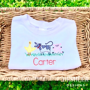 May include: White t-shirt with embroidered farm animals, including a yellow chick, a black and white cow, and a pink pig. The name "Carter" is embroidered in red below the animals.