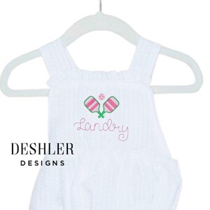Op de afbeelding: Witte seersucker romper met geborduurde pickleball rackets en bal in groen en roze. De naam "Landry" is geborduurd in roze script. Het merk "Deshler Designs" is gedrukt in zwarte blokletters.