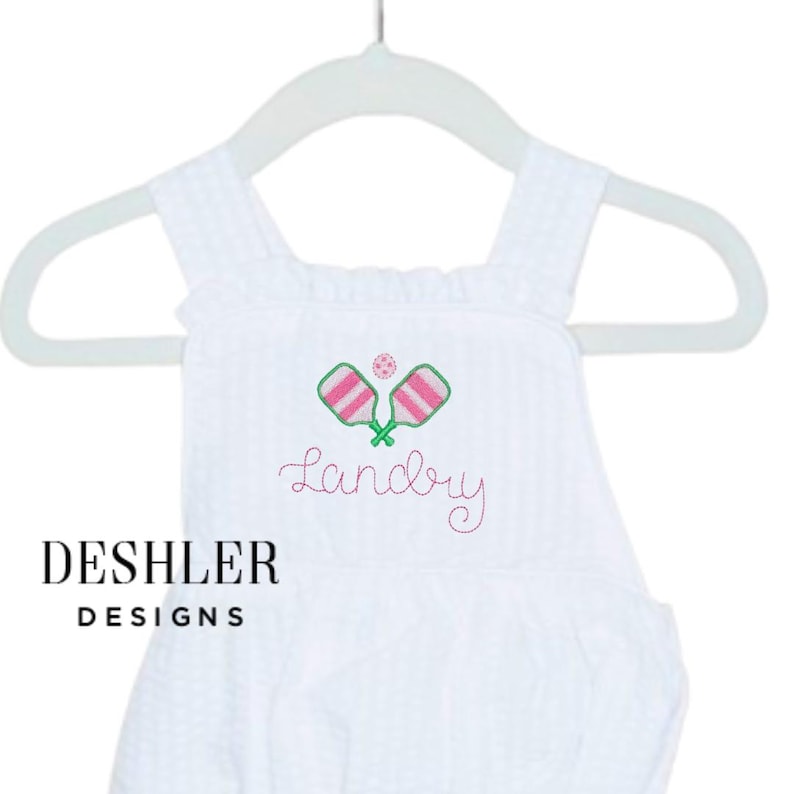 Op de afbeelding: Witte seersucker romper met geborduurde pickleball rackets en bal in groen en roze. De naam "Landry" is geborduurd in roze script. Het merk "Deshler Designs" is gedrukt in zwarte blokletters.