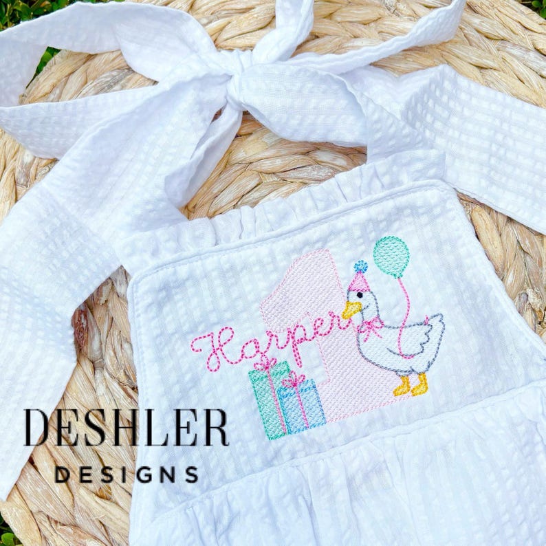 May include: White seersucker bib with a ruffle trim and a large bow. The bib features an embroidered design of a goose wearing a party hat and holding a balloon, with the name "Harper" and the number "1". The text "DESHLER DESIGNS" is printed on the bib.