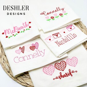 May include: Five white tea towels with embroidered hearts and names. The names are Connelly, McBeath, Nardoillo, Sharife, and Connelly. The towels are folded and stacked on a woven basket.