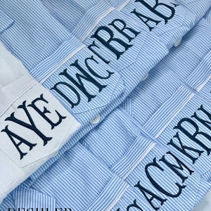 May include: Close-up of several blue and white striped button-down shirts with personalized embroidered initials on the pocket. One white shirt is also visible. The shirts are from Deshler Designs.