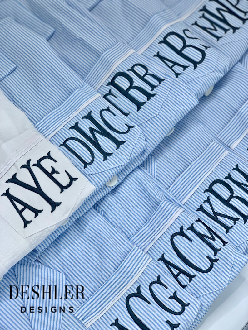 May include: Close-up of several blue and white striped button-down shirts with personalized embroidered initials on the pocket. One white shirt is also visible. The shirts are from Deshler Designs.