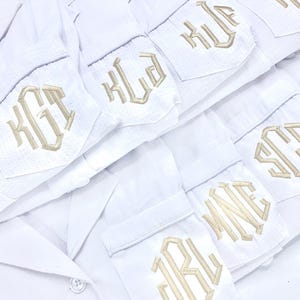 May include: White pajama shirts with embroidered initials in a gold color. The shirts are neatly stacked, with the embroidered initials visible on the pocket. The text "DESHLER DESIGNS" is at the bottom of the image.