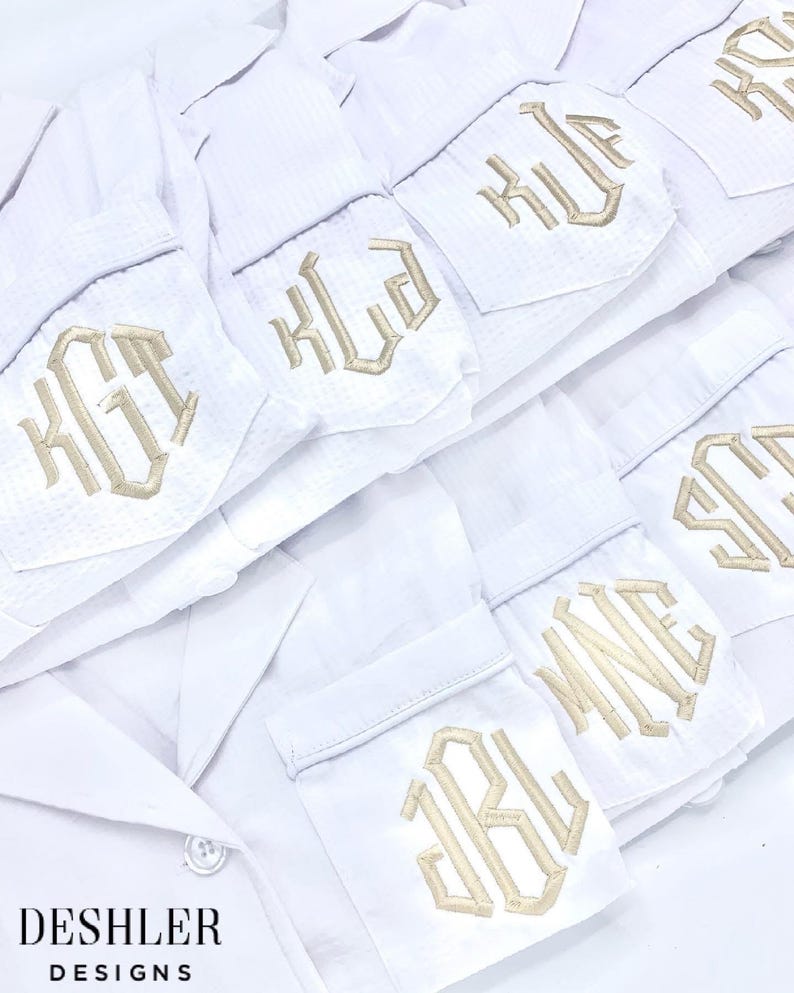 May include: White pajama shirts with embroidered initials in a gold color. The shirts are neatly stacked, with the embroidered initials visible on the pocket. The text "DESHLER DESIGNS" is at the bottom of the image.