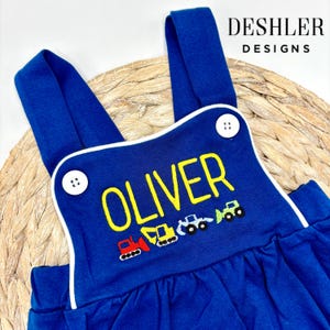 May include: Blue overalls with white piping and a yellow embroidered name "OLIVER" and a row of colorful embroidered construction vehicles.
