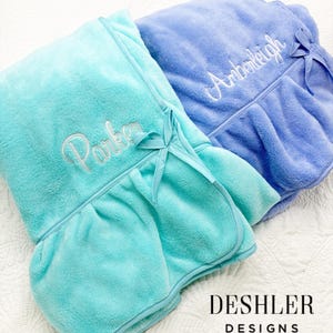 May include: Two plush, personalized spa wraps in shades of teal and periwinkle. Each wrap features a ruffled hem, a matching bow, and embroidered names in elegant script. The wraps are from Deshler Designs.