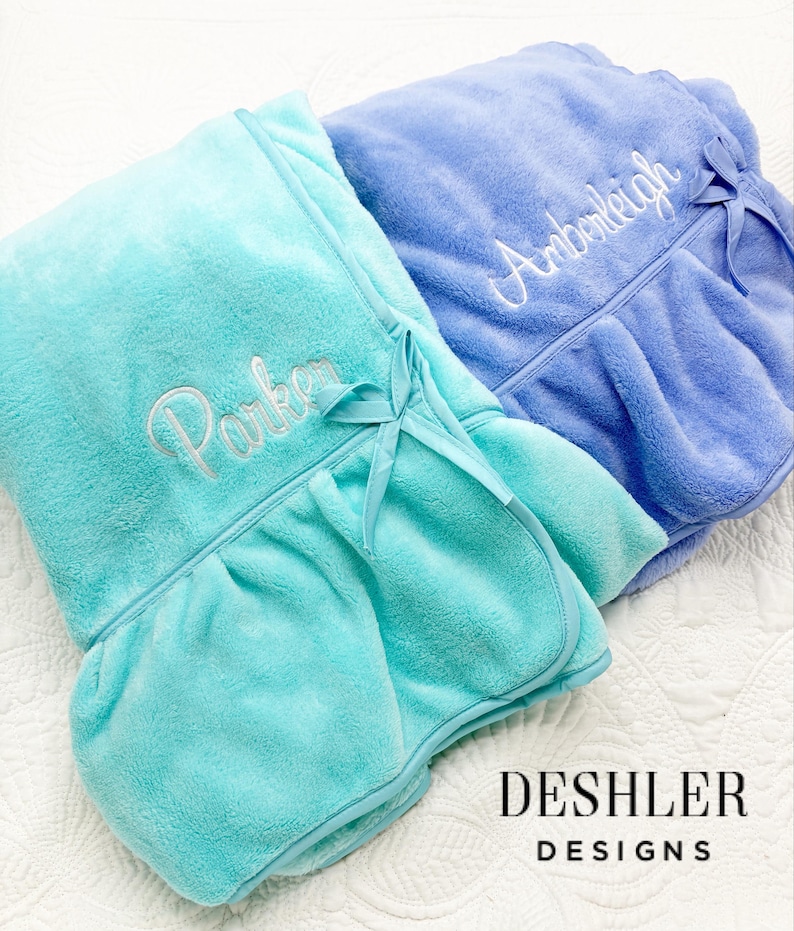 May include: Two plush, personalized spa wraps in shades of teal and periwinkle. Each wrap features a ruffled hem, a matching bow, and embroidered names in elegant script. The wraps are from Deshler Designs.