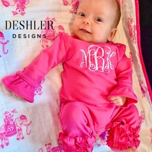 May include: A pink baby onesie with ruffled sleeves and legs. The onesie has a white embroidered monogram "MBK" on the front. The onesie is lying on a white blanket with a pink elephant print.