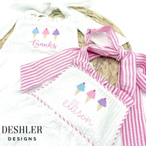 May include: Two white sleeveless dresses with pink and white striped straps. The dresses feature embroidered ice cream cones and the names "Banks" and "ellison".