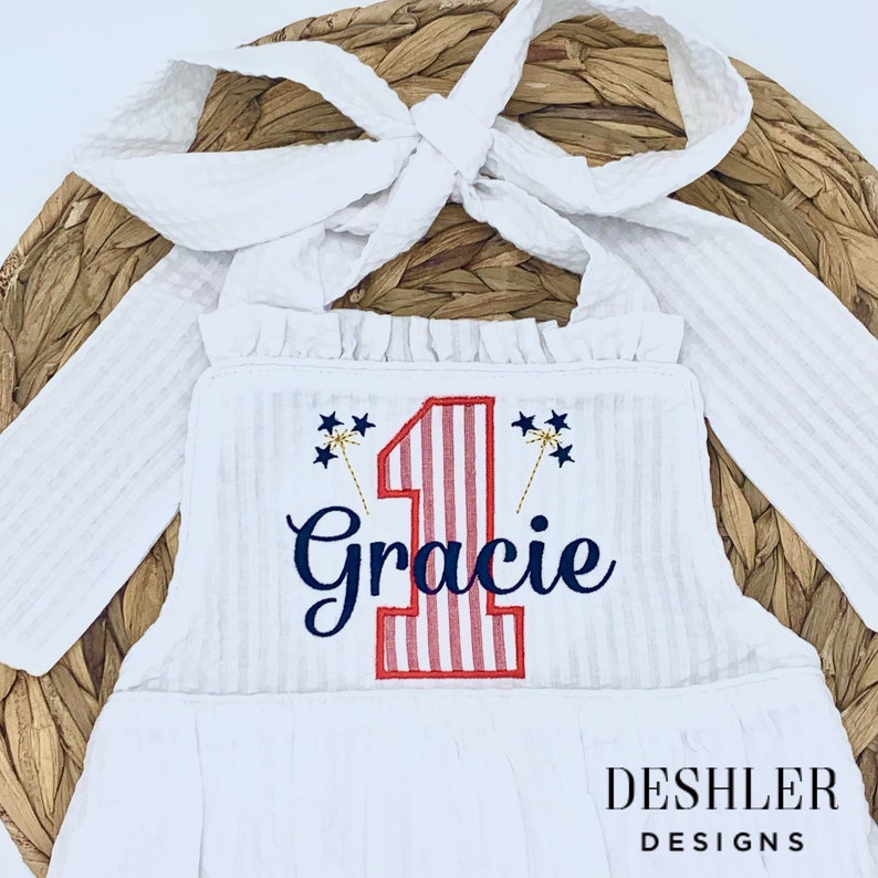 May include: White sleeveless dress with red and white striped number one and the name Gracie embroidered in navy blue. The dress has a white bow tie and is decorated with gold glitter fireworks.