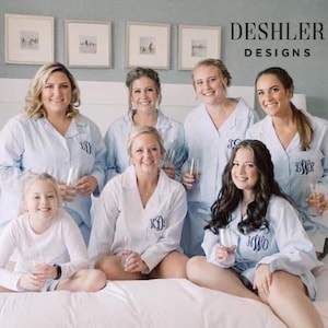 May include: A group of women wearing light blue monogrammed button-down shirts and white shirts, some holding glasses. The text "DESHLER DESIGNS" is visible in the upper right corner. The women are sitting on a bed.