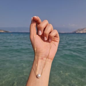 May include: A delicate gold chain necklace with a single, large, white pearl pendant. The necklace is held up against a backdrop of the ocean and a clear blue sky. The chain appears to be approximately 20 inches long.