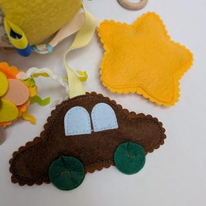 Op de afbeelding: Handgemaakte baby mobile met een bruine vilten auto met blauwe ramen en groene wielen, een gele ster en andere kleurrijke vilten vormen. Inclusief een houten ring en linten.