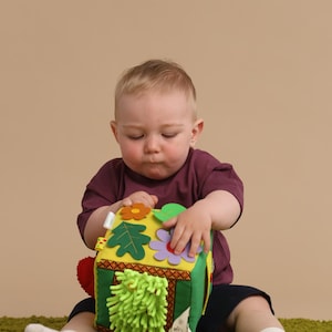 Può includere: Un cubo attività in tessuto morbido e colorato per bambini. Il cubo presenta varie trame, forme e colori, tra cui fiori, foglie e una frangia verde. Il giocattolo è progettato per stimolare lo sviluppo sensoriale.