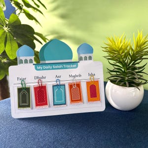 May include: A white rectangular My Daily Salah Tracker board featuring a blue mosque illustration at the top. It includes five prayer labels: Fajar, Dhuhr, Asr, Maghrib, and Isha, each with a small colorful prayer rug card attached by a paperclip.
