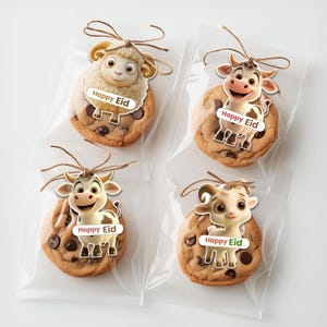 Puede incluir: Cuatro galletas con chispas de chocolate en bolsas transparentes, cada una adornada con un animal de dibujos animados y una etiqueta "Happy Eid". Los animales incluyen una oveja, una vaca y una cabra, atados con hilo.