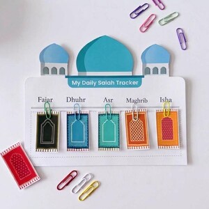 May include: A white My Daily Salah Tracker board shaped like a mosque with a blue dome. It features five columns labeled Fajar, Dhuhr, Asr, Maghrib, and Isha, each with a small rectangular prayer rug card attached by a colorful paper clip.