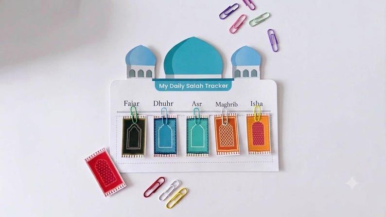 May include: A white My Daily Salah Tracker board shaped like a mosque with a blue dome. It features five columns labeled Fajar, Dhuhr, Asr, Maghrib, and Isha, each with a small rectangular prayer rug card attached by a colorful paper clip.