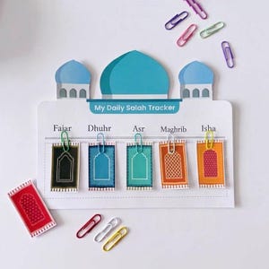 May include: A white My Daily Salah Tracker board shaped like a mosque with blue domes. It features five prayer names: Fajar, Dhuhr, Asr, Maghrib, and Isha. Five small rectangular prayer rug cards are attached with colorful paper clips.