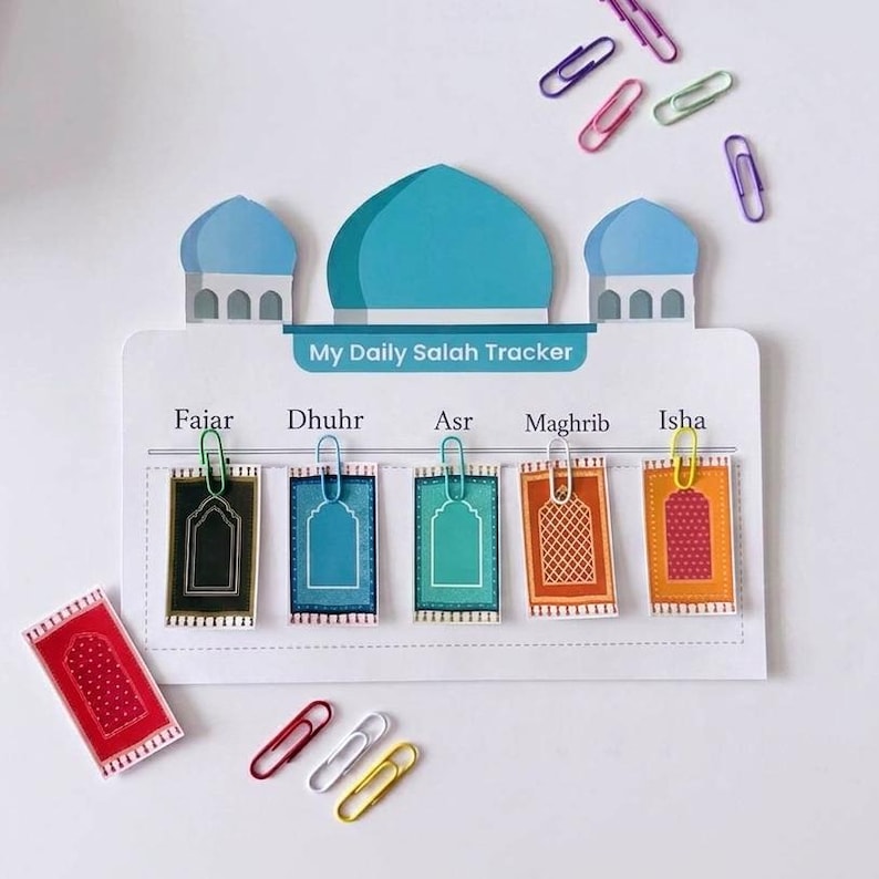 May include: A white My Daily Salah Tracker board shaped like a mosque with blue domes. It features five prayer names: Fajar, Dhuhr, Asr, Maghrib, and Isha. Five small rectangular prayer rug cards are attached with colorful paper clips.
