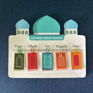 May include: A white My Daily Salah Tracker board featuring a mosque silhouette with three teal and blue domes. Five small rectangular prayer rug cards in green, red, teal, orange, and pink are attached with colorful paper clips below prayer names.