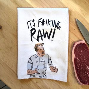 May include: White kitchen towel with the text "IT'S F#IKING RAW!" and a cartoon image of a chef. A raw steak and a knife are on a wooden surface next to the towel.