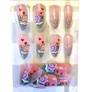 May include: A set of artificial nails with a pink and nude gradient design. Each nail features a cartoon snail with hearts and bows. The nails are packaged in a clear plastic container.