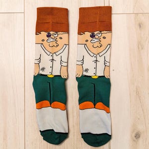 May include: A pair of novelty socks featuring a cartoon character design. The socks have a brown cuff, a tan face, a white shirt, green pants, and orange shoes. The socks are displayed on a light wood surface.