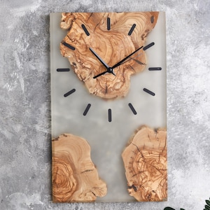 以下が含まれることがあります： 明るい灰色の樹脂の背景に木のスライスが埋め込まれた長方形の壁掛け時計。時計には黒い針と黒い時間マーカーがあります。木材は自然な薄茶色で、木目が見えます。