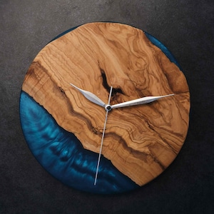May include: A circular wall clock featuring a natural light brown wood grain slab with a prominent dark knot, contrasted by a deep blue translucent resin section. The clock includes three thin, silver-colored metal hands centered on the face.