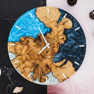 May include: A round wooden clock face with blue and black resin accents. The clock face has a natural wood grain pattern and white clock hands.